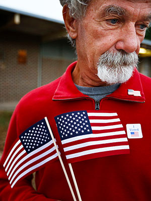 Man wearing an "I voted" sticker holding two American flags