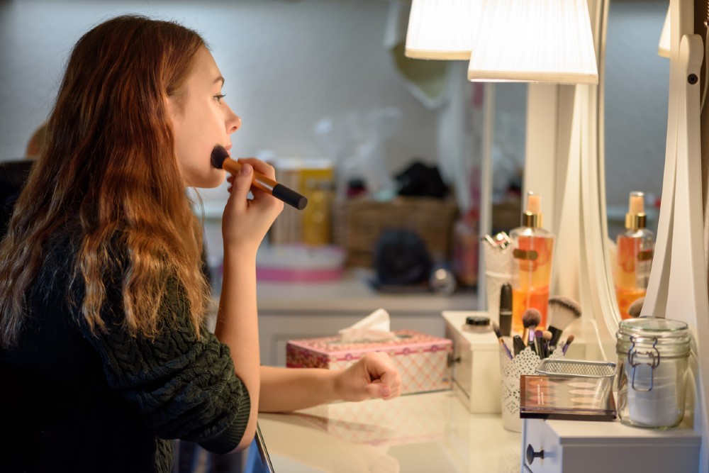 A woman applying foundation for dry skin onto her cheeks with a brush as she looks into a mirror