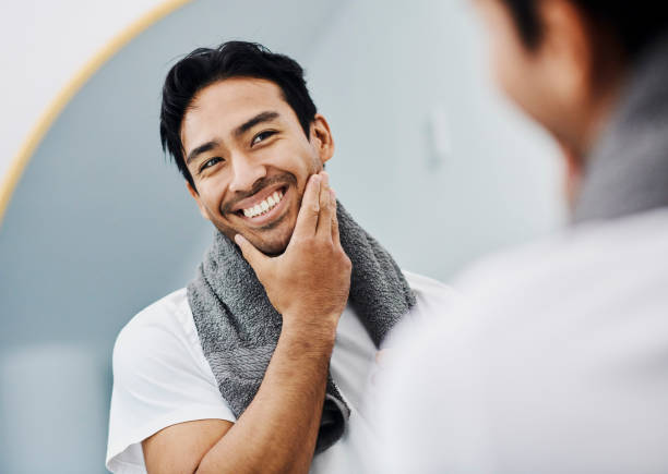 Man happy about face skin care product for healthy body and facial skincare for guys. Handsome young male looking in the mirror with toothy smile, touching his beard satisfied with his shaving cream