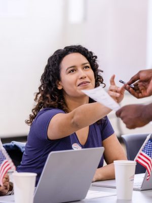 Two cheerful female volunteers help the unrecognizable young adult with his ballot for the election.